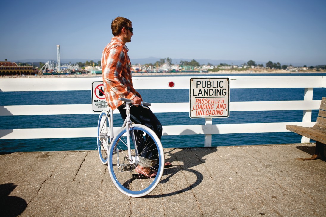 Man in orange plaid shirt riding Globe Roll bike on Santa Cruz pier with ocean and amusement park in background