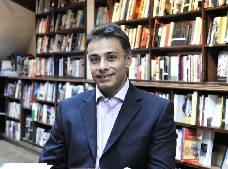 Man in navy blazer seated at table with books in Daunt Books bookshop with shelves behind.