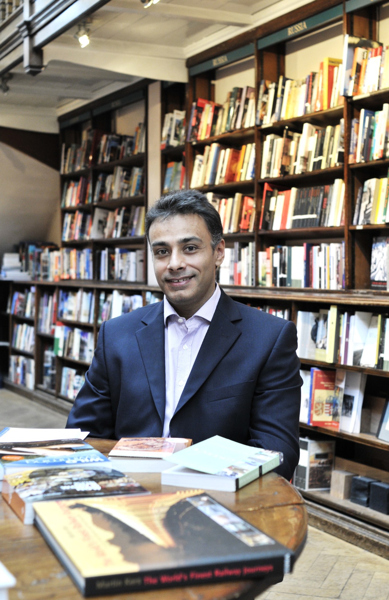 Man in navy blazer seated at table with books in Daunt Books bookshop with shelves behind.