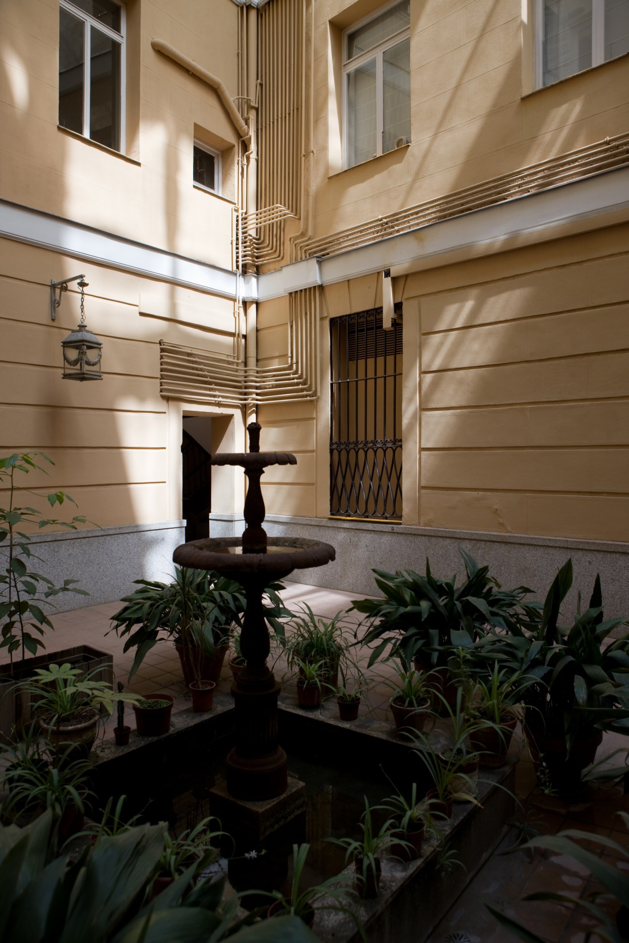 Sunlit courtyard of a Malasaña apartment building with cream-colored walls, decorative metal piping, potted plants, and a central fountain.
