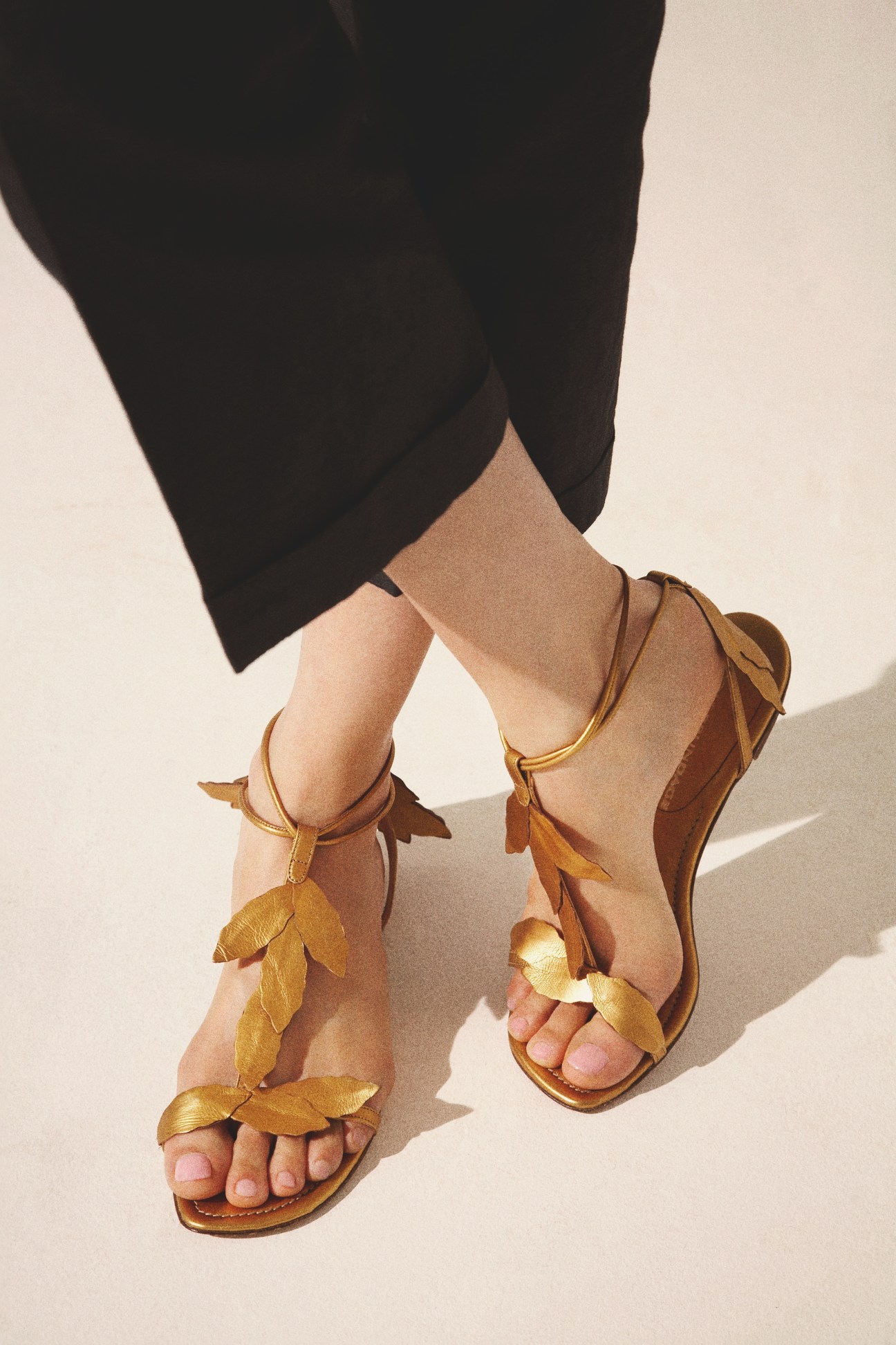 Woman wearing brown leather sandals with gold leaf embellishments and black trousers.