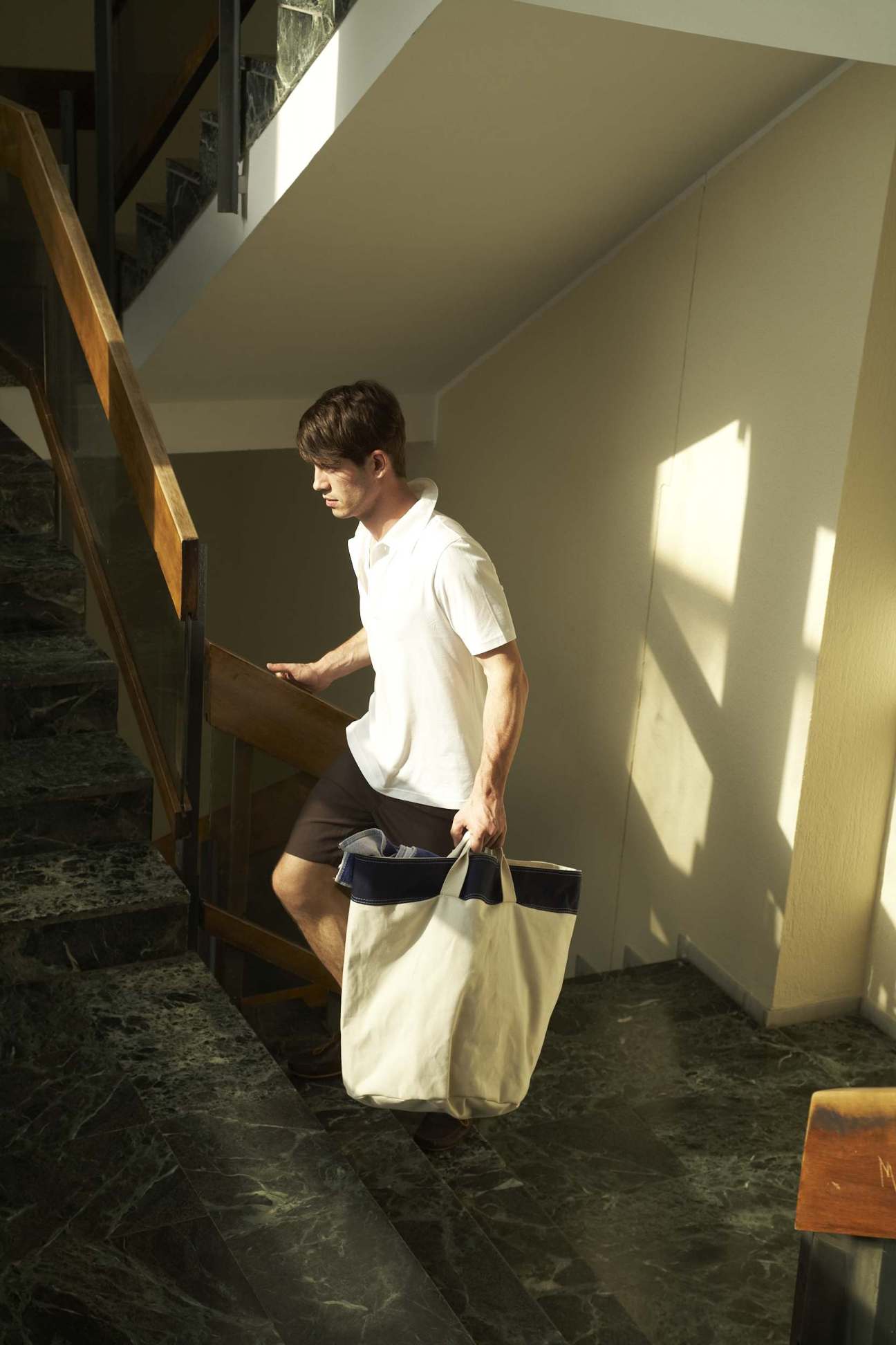 Man in white polo shirt and cream linen shorts walking through a sunlit corridor with a navy striped tote bag.