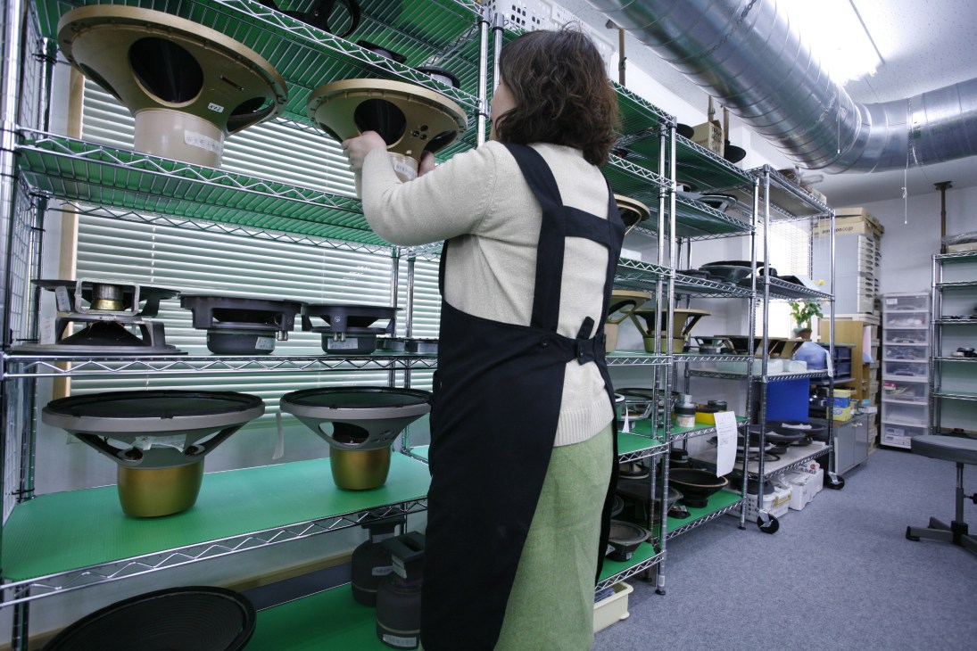 A technician reaches toward vintage speakers on metal shelving at Audiolab's repair workshop in Japan.