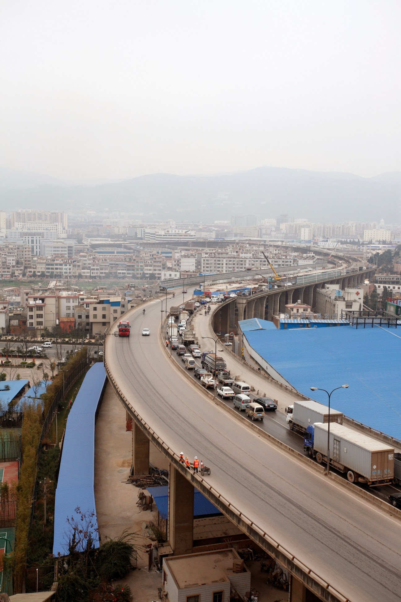 Elevated highway viaduct with trucks and construction equipment overlooking Kunming's residential cityscape and mountains.