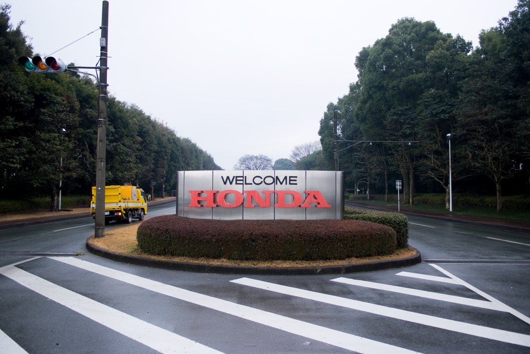 Welcome sign at Honda entrance with large red letters on white panel beside a tree-lined road.