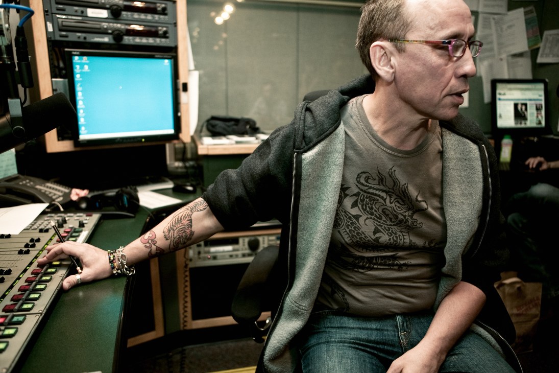 Nic Harcourt sits at a KCRW radio station control board with mixing equipment and computer monitors in the studio.