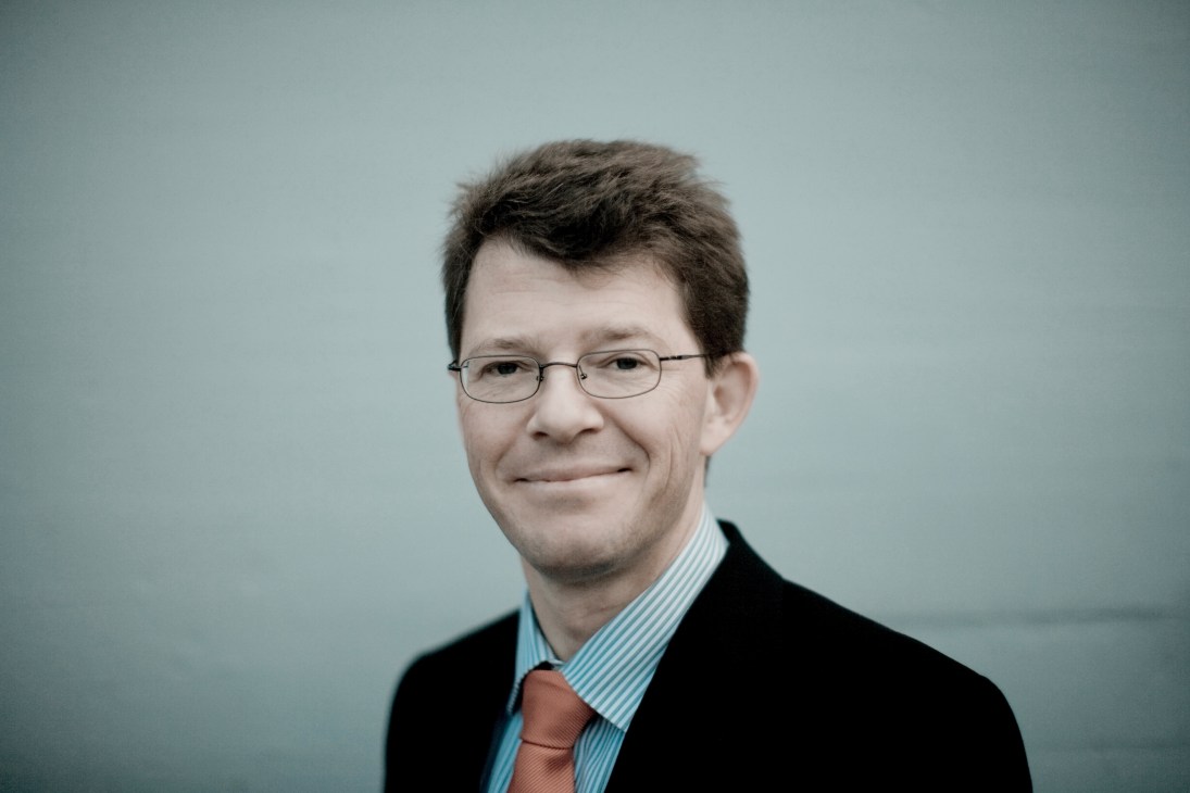 Portrait of Jonas Hughes, Schindler's communications director, wearing glasses and a dark blazer with striped shirt and tie.