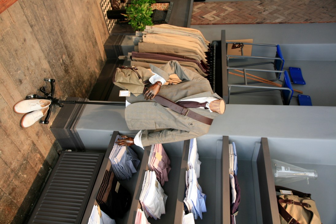 Man wearing beige suit and burgundy belt stands in Oliver Spencer store surrounded by organized clothing displays.