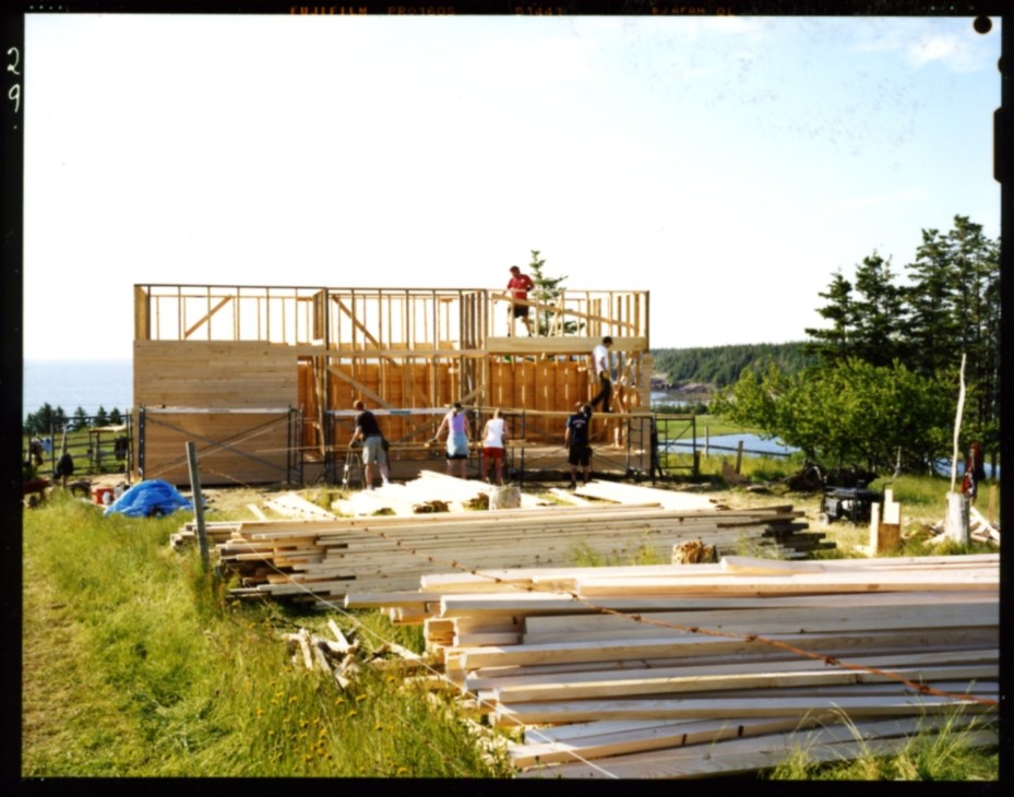 Construction site of wooden building frame with students and workers on scaffolding overlooking coastal landscape.