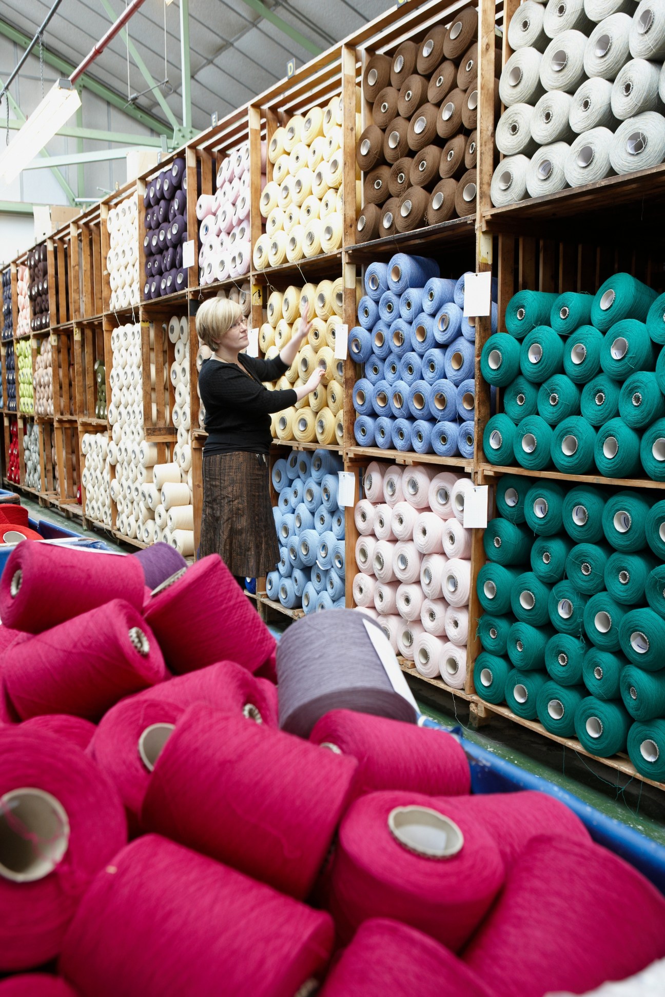 Woman examining colorful cashmere yarn spools organized on wooden shelves at Johnstons factory in Scotland.