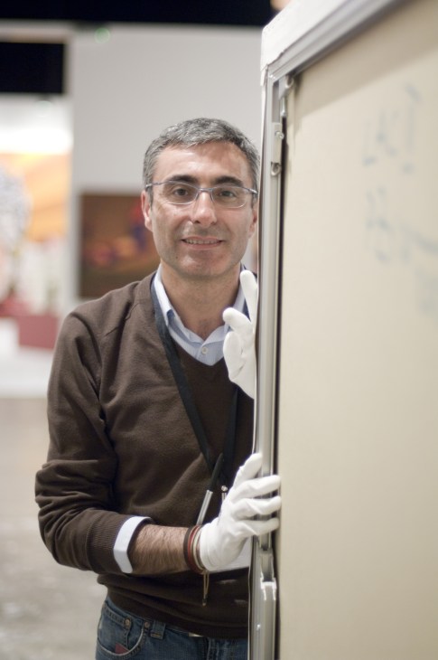 A man in white gloves and brown sweater holds a gallery door frame during Art Dubai setup.