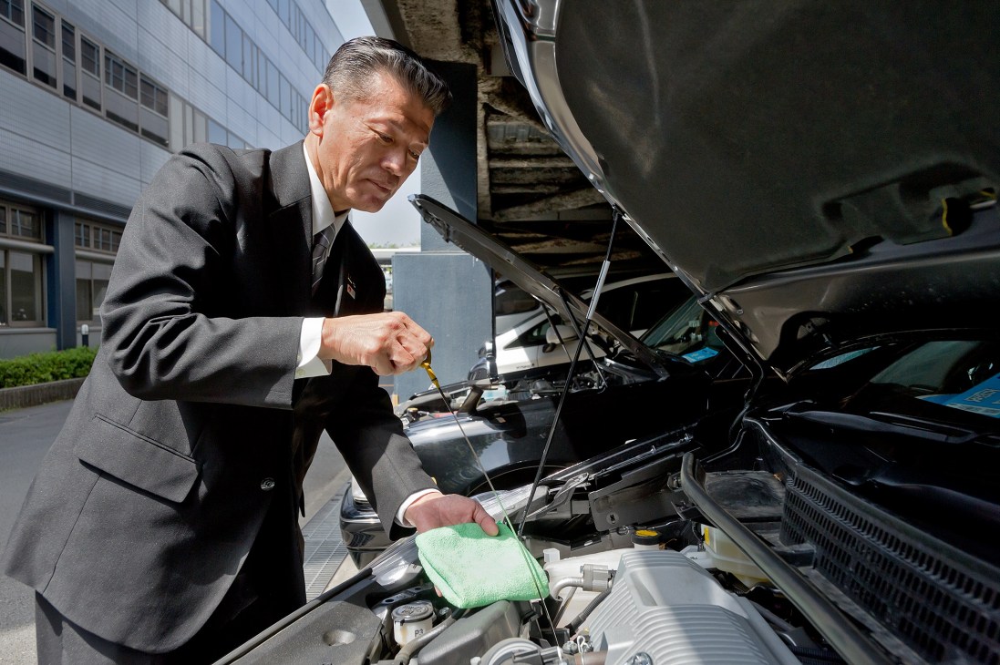 A taxi driver in formal attire performs routine maintenance checks on a vehicle engine at the depot.