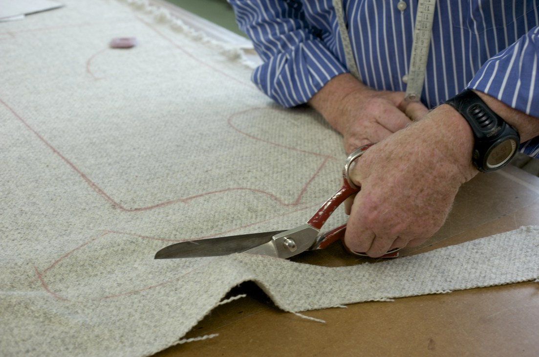 A tailor's hands marking and cutting cream-colored lambswool fabric with chalk and scissors.