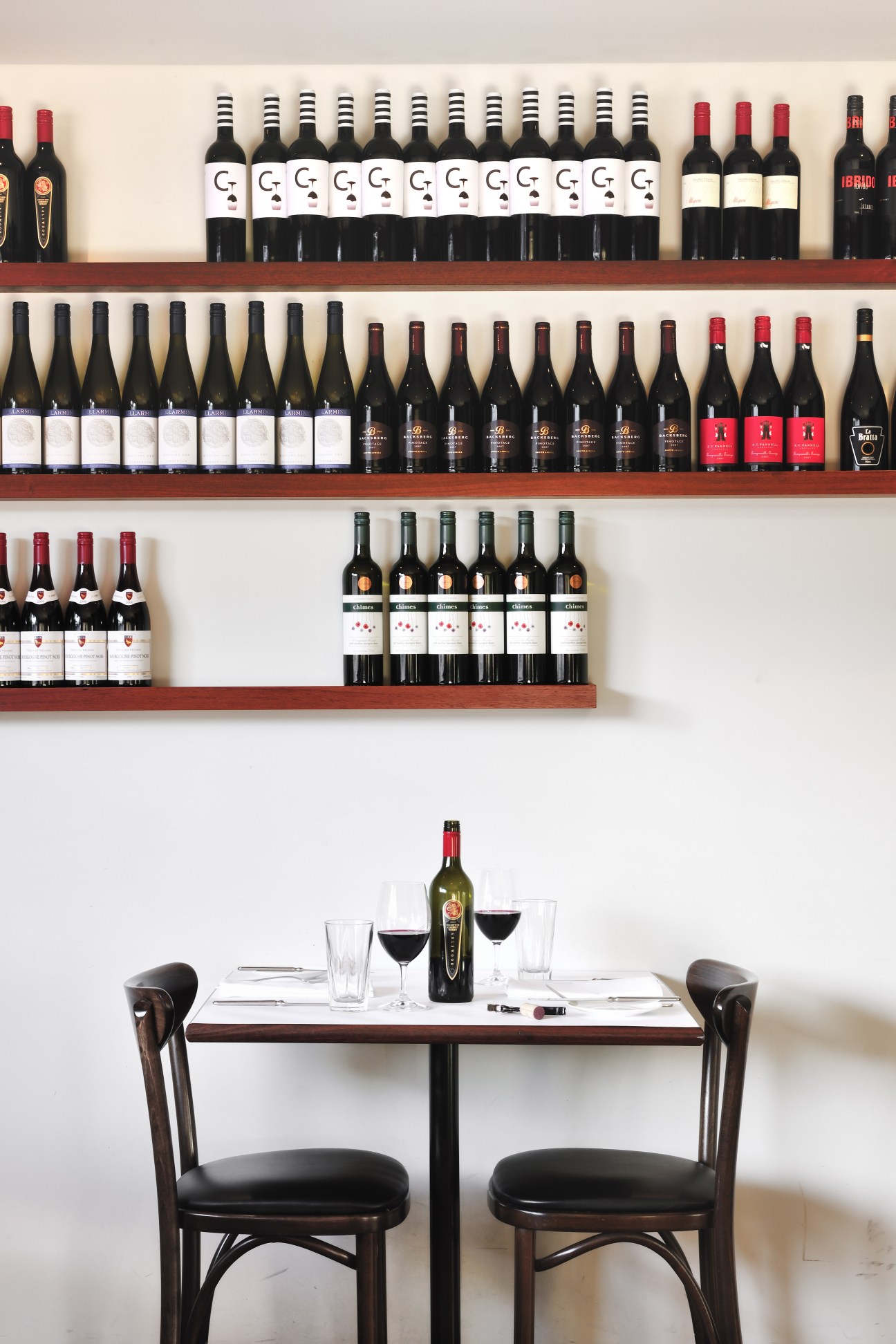 Wine bottles displayed on wooden shelves above a dining table set with glasses and a bottle.