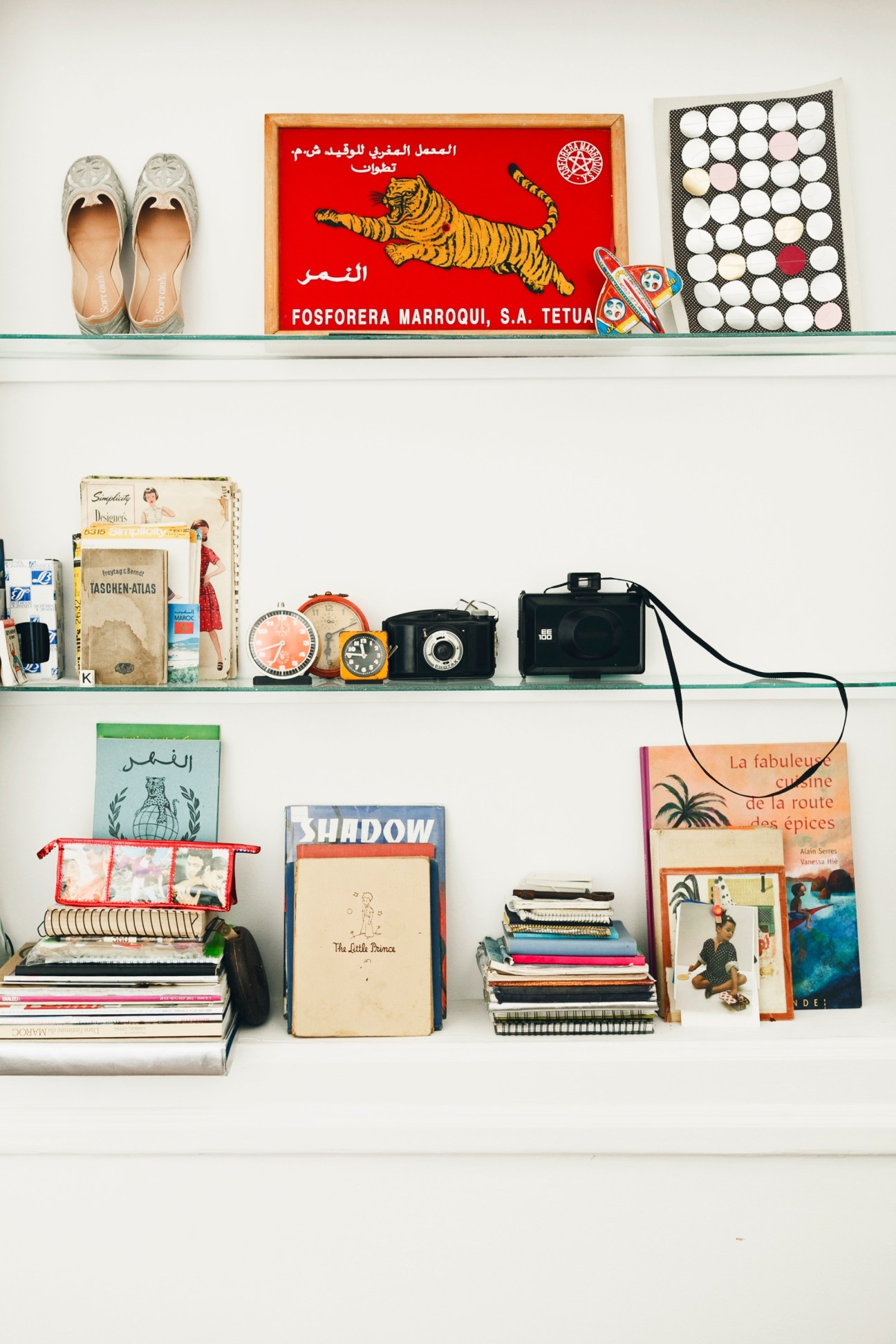White shelves displaying vintage cameras, books, shoes, and decorative objects including a Moroccan matchbox with tiger design.