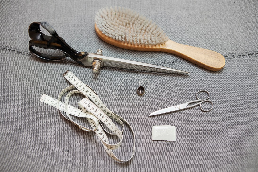 Tailoring tools arranged on grey fabric including scissors, measuring tape, brush, and needle.
