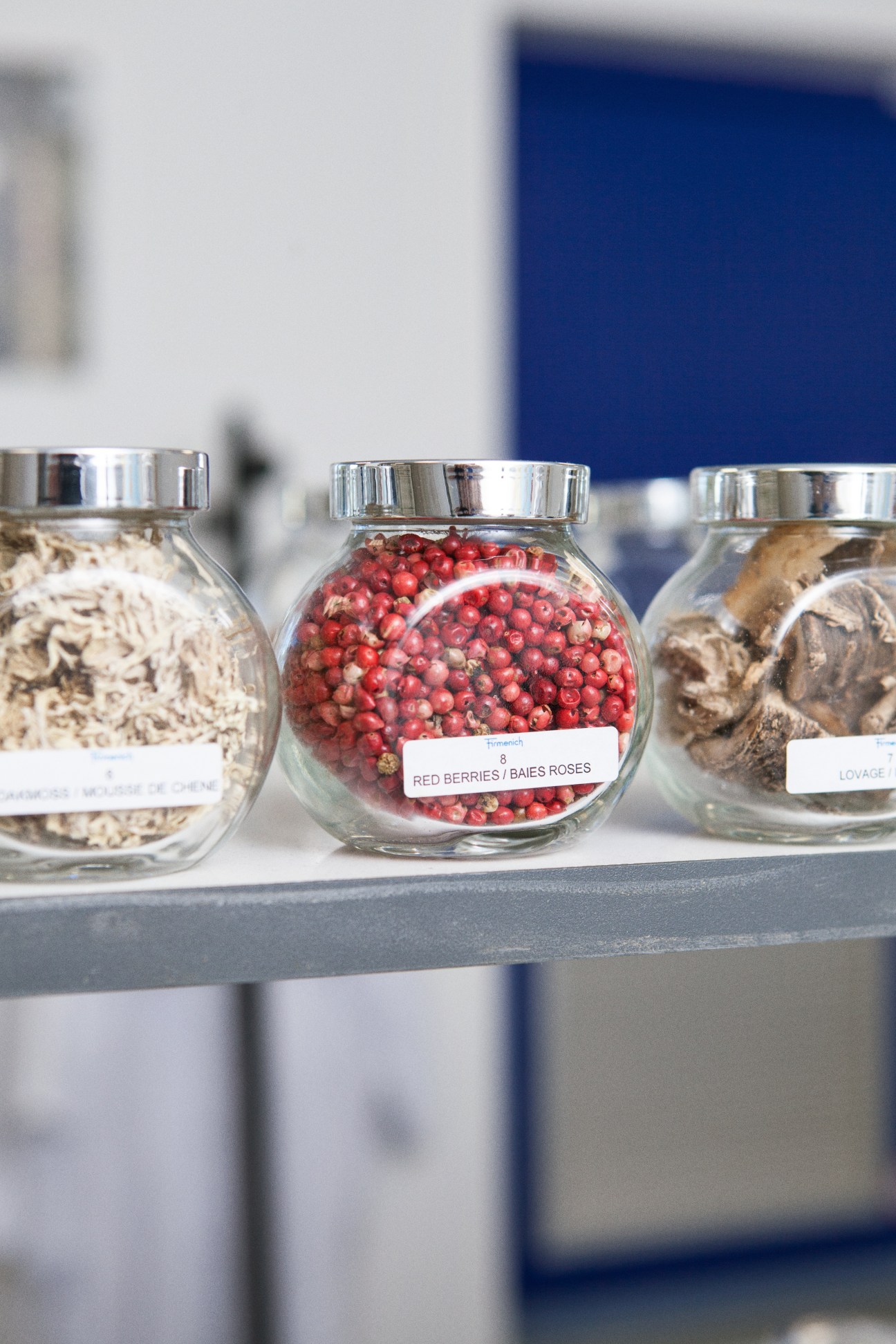 Glass jars containing natural perfume ingredients including red berries and dried botanicals labeled on a shelf.