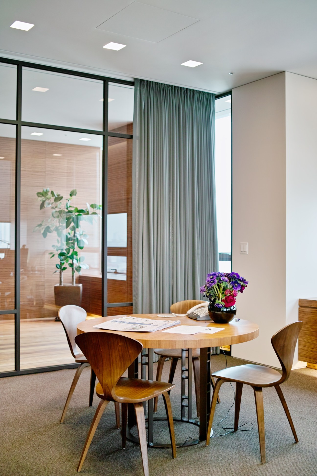 Modern office meeting room with oval wooden table, mid-century chairs, glass partitions, and green curtains.