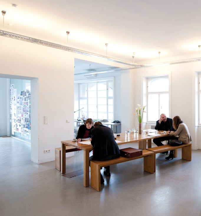 Interior of Gregor Eichinger's minimalist architecture offices with wooden tables, white walls, and people working.