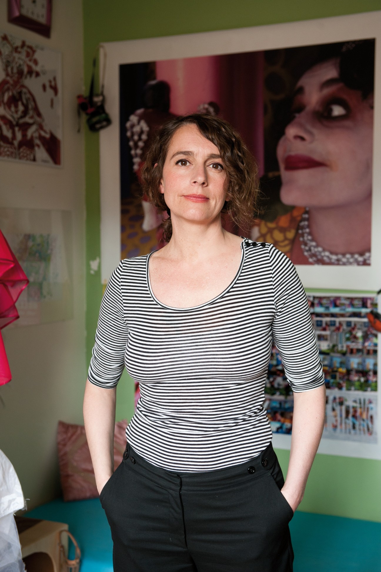 Performance artist Bella Angora stands in a colorful studio space with vibrant artwork on the walls.