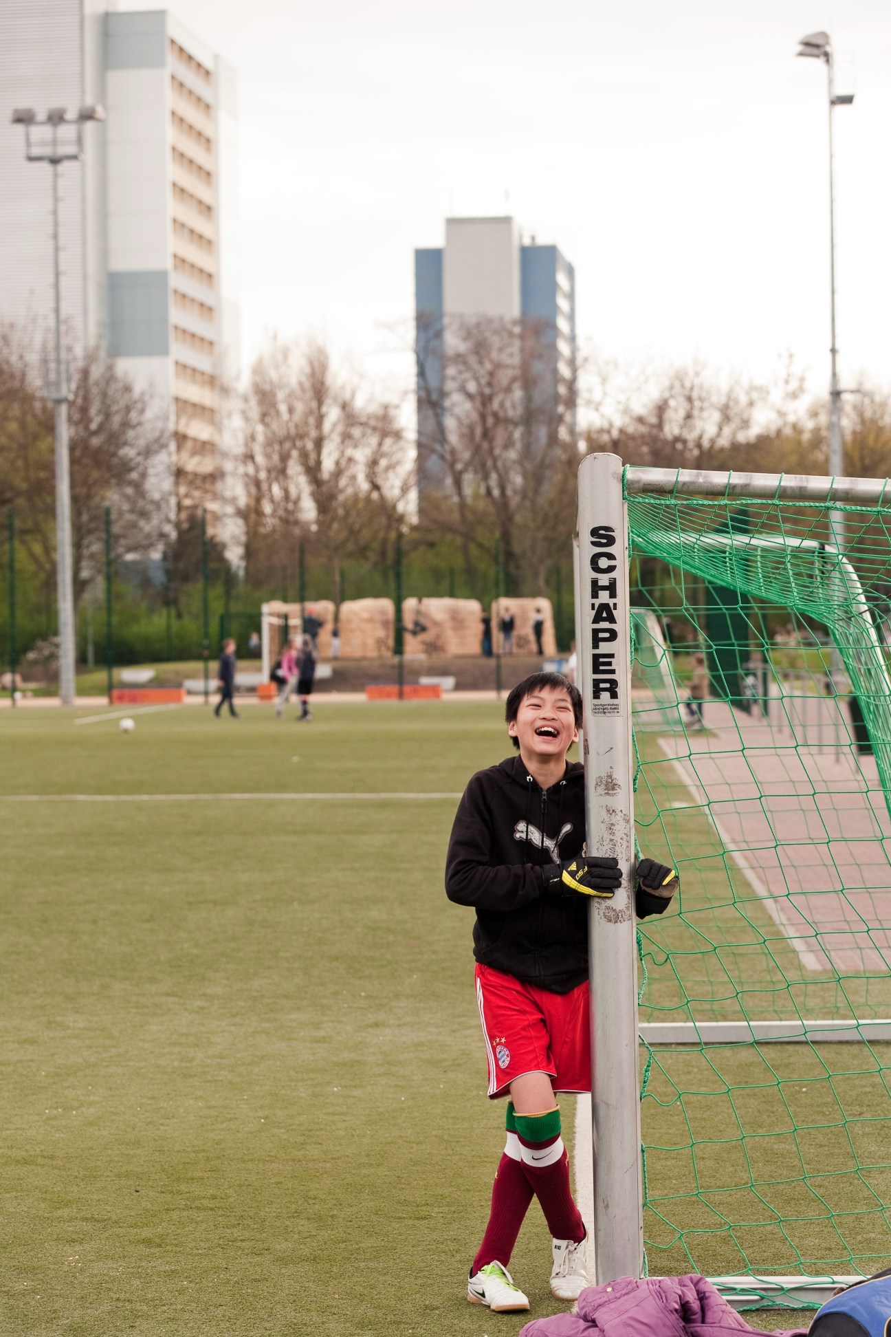 Young boy laughing while leaning against a green soccer goal post on a field with apartment buildings in the background.