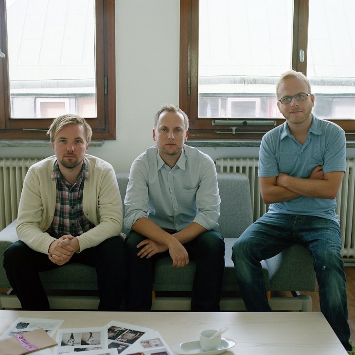 Three men from Next Century Modern sitting on a couch in an office with large windows.