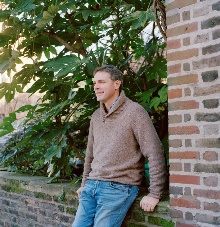 Man in beige sweater and jeans standing against brick wall with climbing ivy.