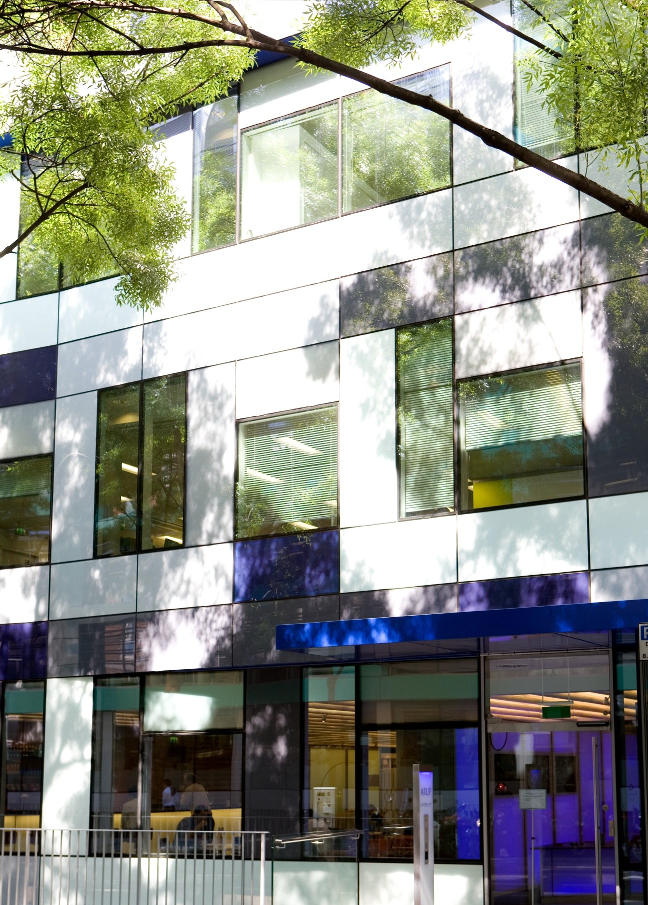 Modern office building exterior with white and blue panel facade, large windows, and tree branches overhead.