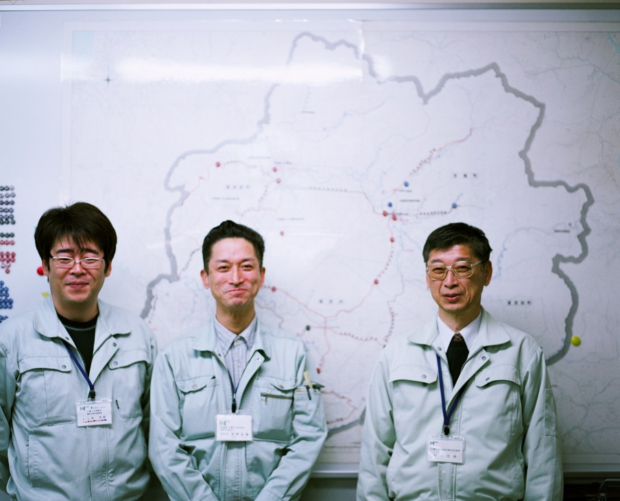 Three Japanese government workers in light-colored uniforms stand in front of a large map at the local government office.