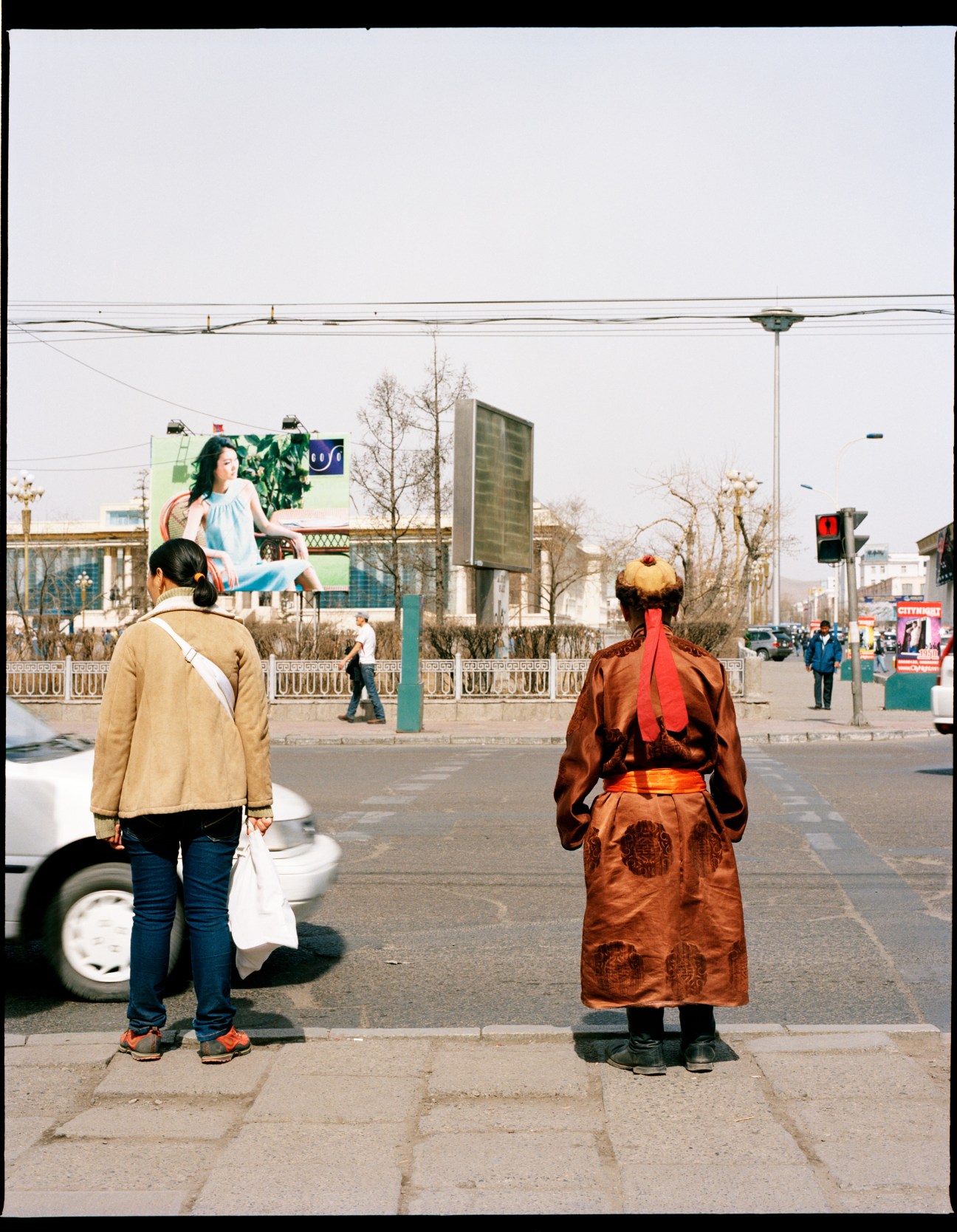 Two women in Ulan Bator wearing contrasting styles: one in a beige jacket and jeans, the other in a traditional brown Mongolian dress with red sash.