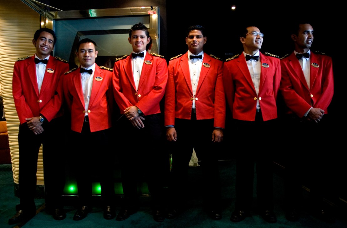 Six bellhops in red uniforms with gold trim and bow ties stand in a line.