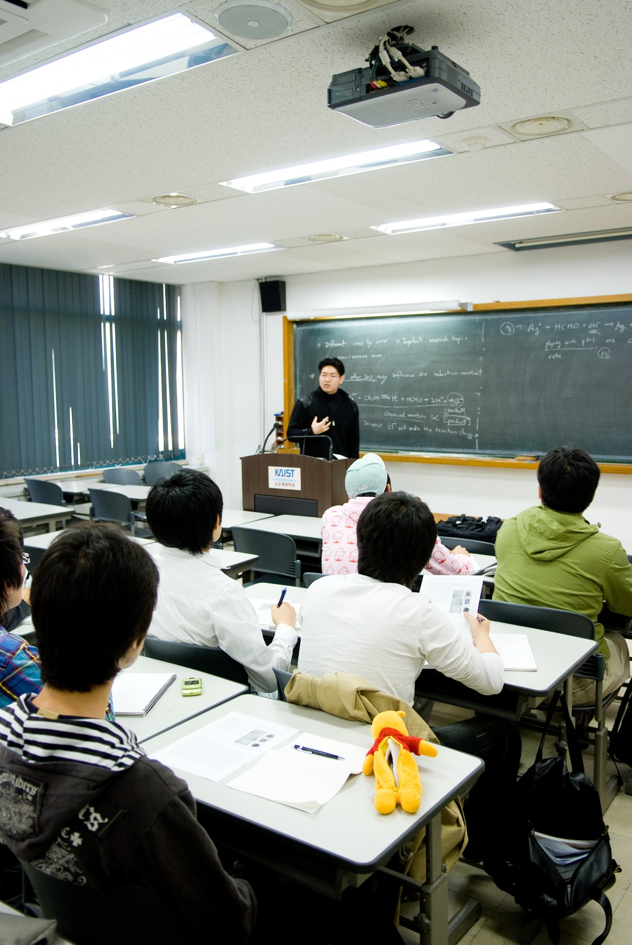 KAIST students sit at desks during a class taught by an instructor standing at a podium with a blackboard of equations behind her.