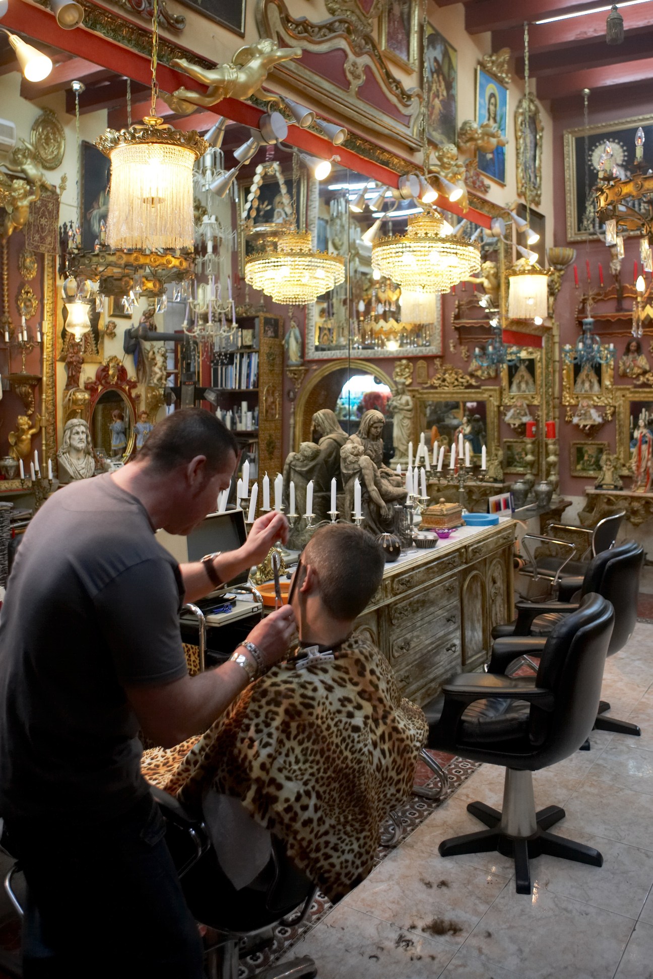 Barber cuts customer's hair in ornately decorated shop with religious statues and golden chandeliers.