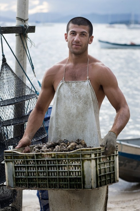 Laelcio Queiroz, an oyster farmer, holds a crate of oysters on a boat with the sea behind him.
