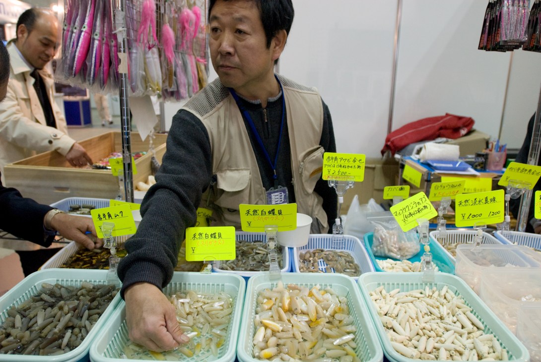 Man displaying various types of fish bait in containers at a boat show exhibit.