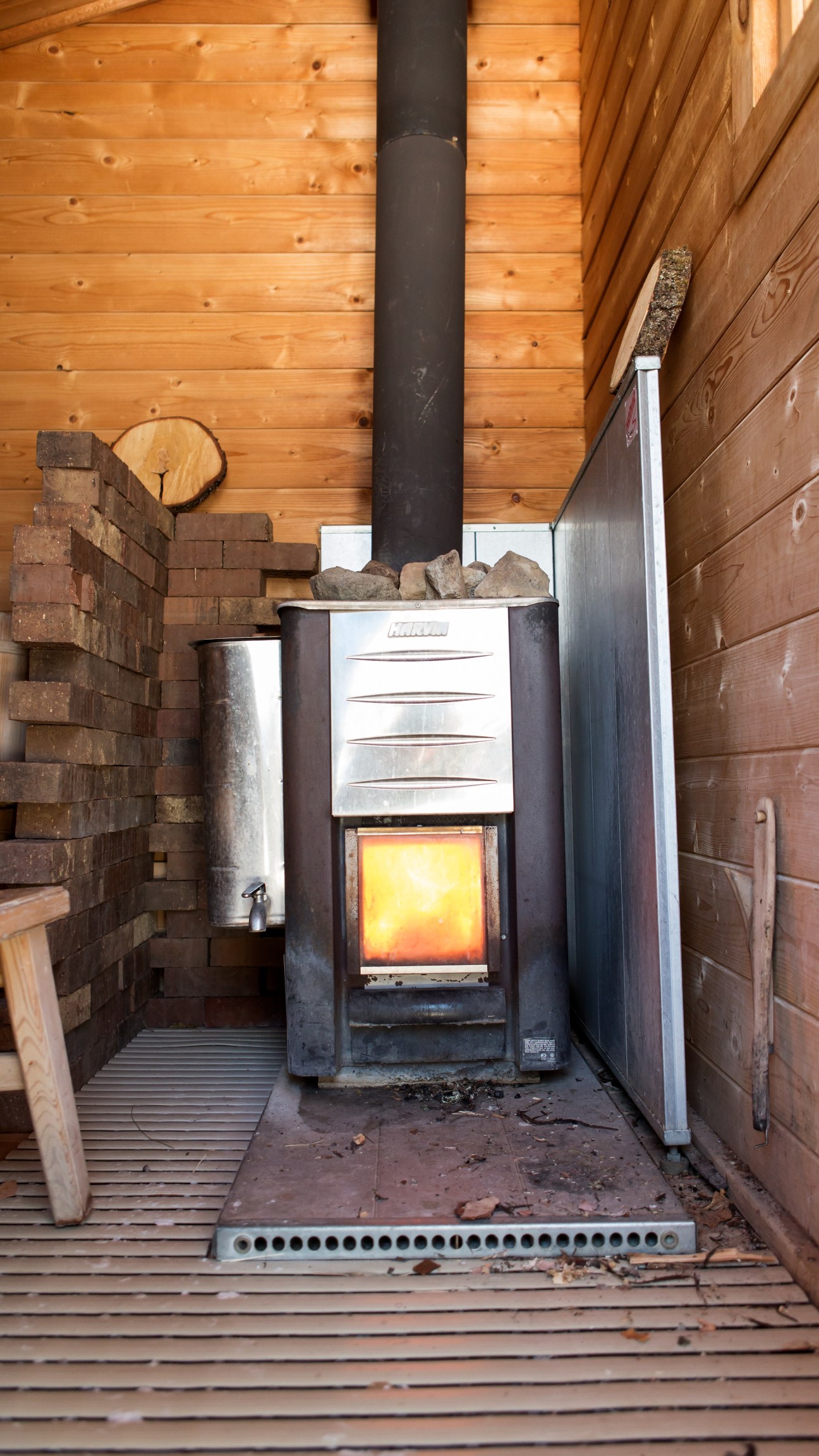 Wood-burning stove glowing with fire inside a traditional sauna with wooden walls and birch-bark brush hanging on the wall.