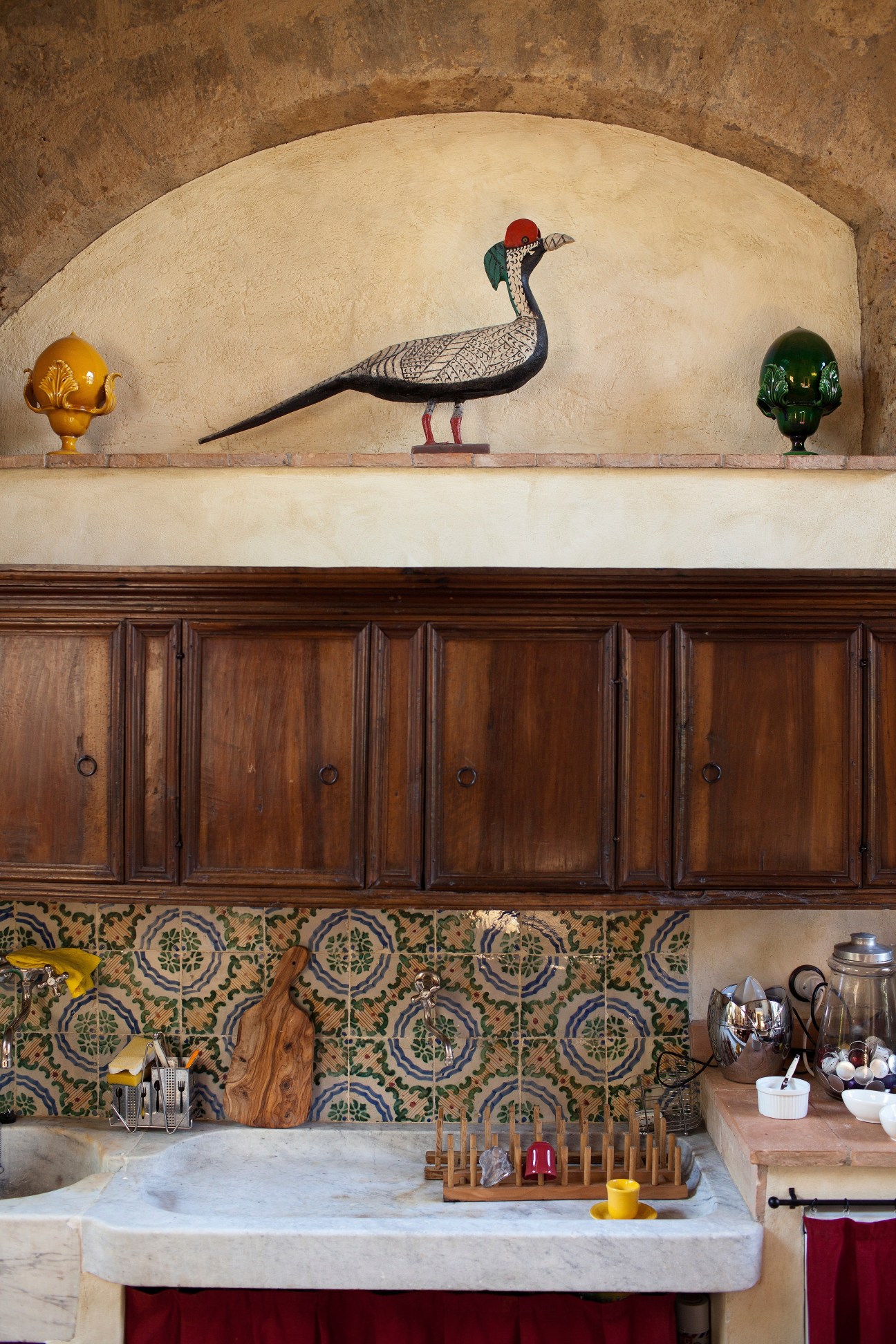 Kitchen niche with decorative bird sculpture and golden ornaments above wooden cabinetry and patterned tile backsplash.