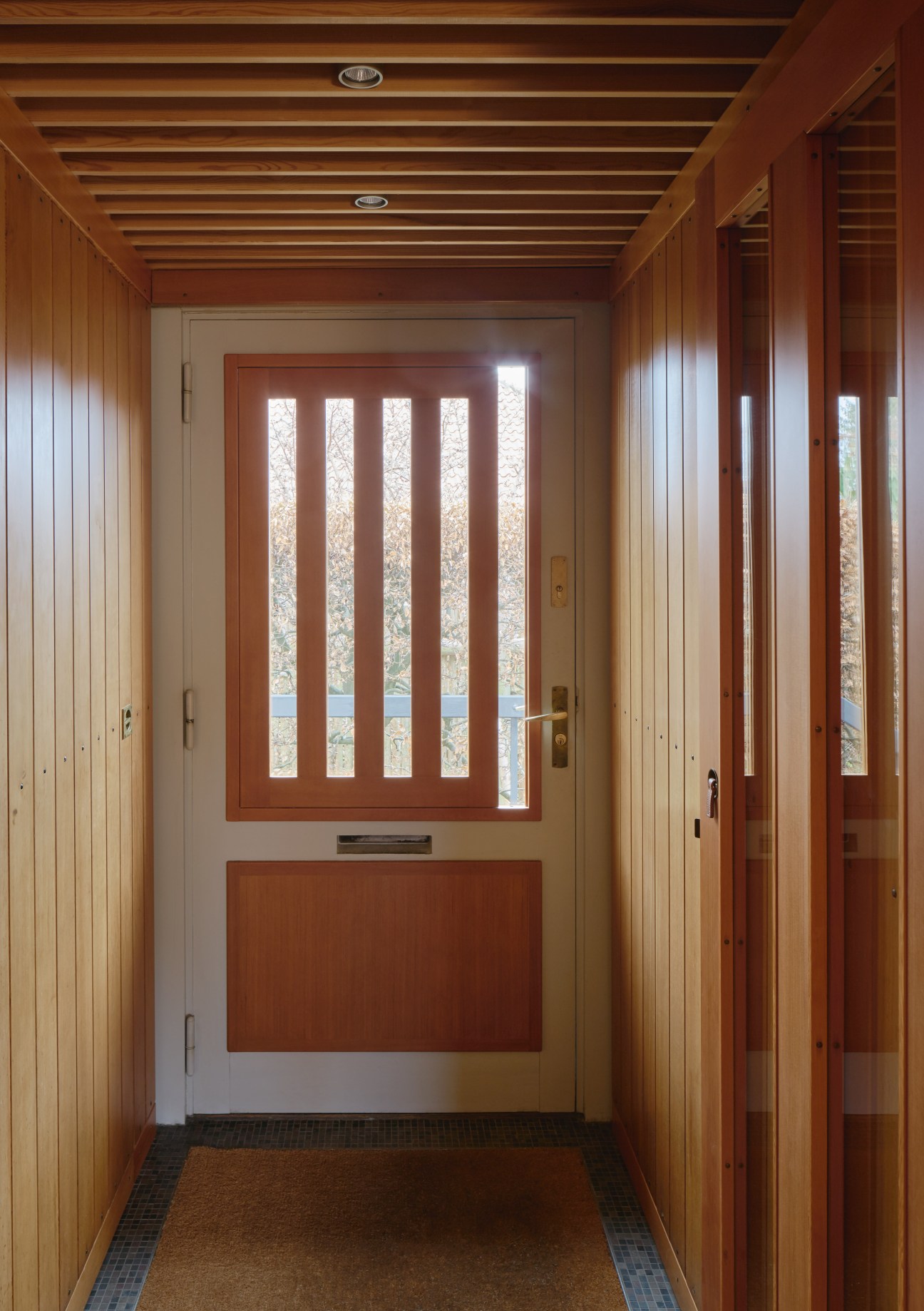 A modern entryway with wooden panelled walls and a red door featuring vertical glass panes with movable wooden shades.