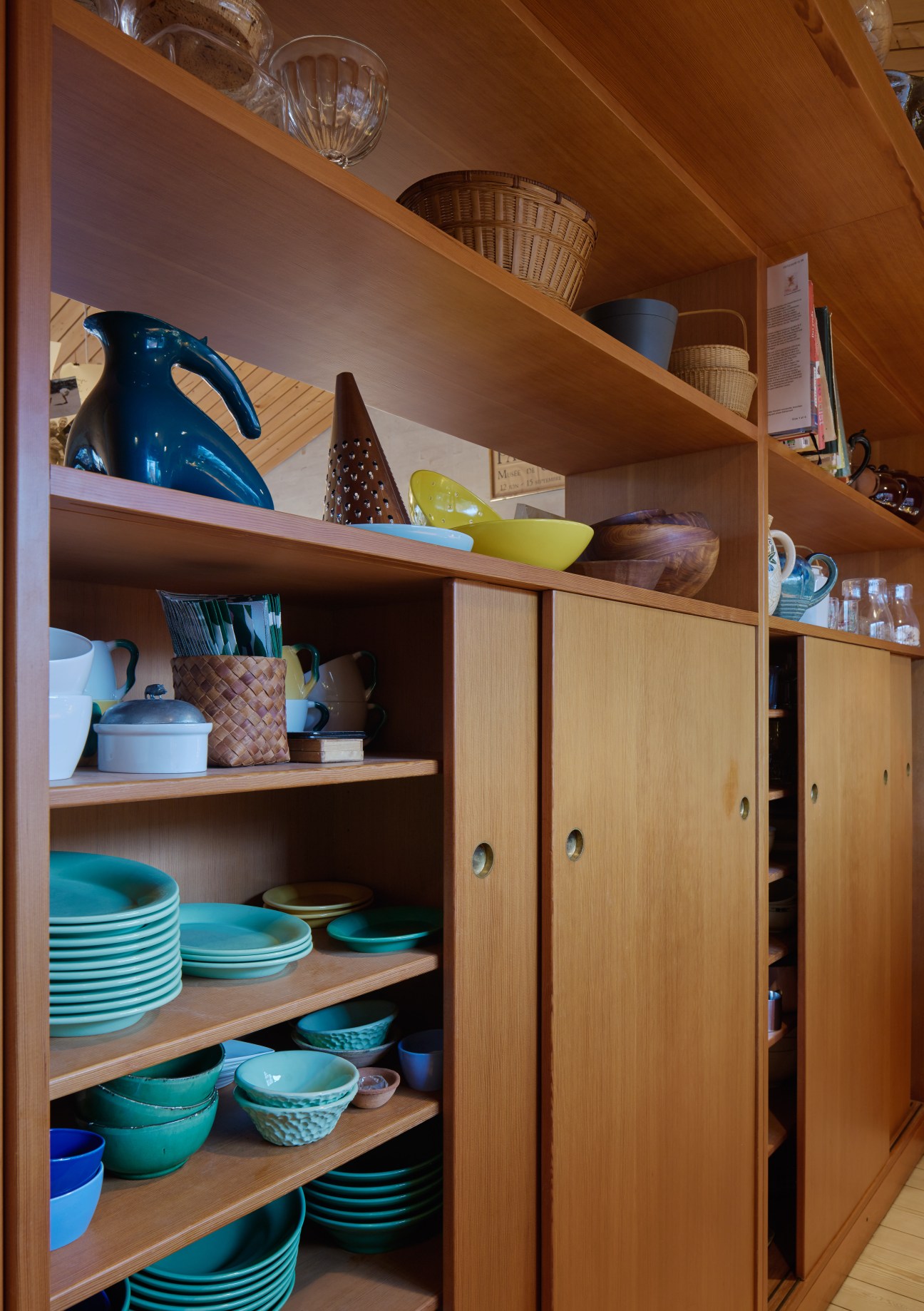Open wooden shelving unit displaying colorful bowls, baskets, and dinnerware with hanging light fixtures above.