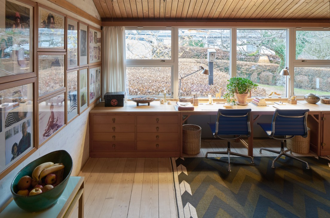 A bright home office and family room with wooden desks, blue chairs, and large windows overlooking a garden.