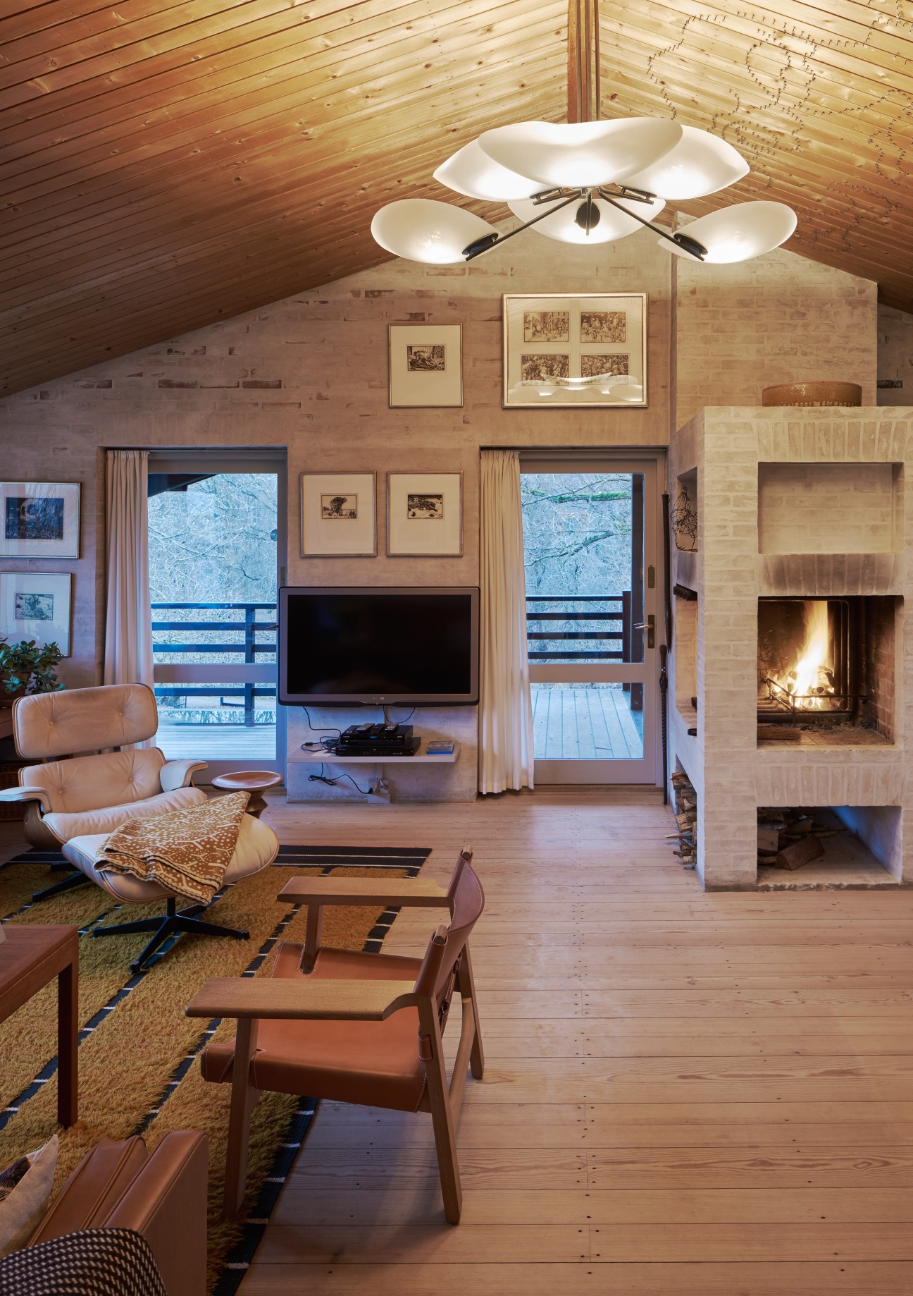 Modern living room with wooden ceiling, fireplace, mid-century furniture, and glass doors opening to snowy landscape.