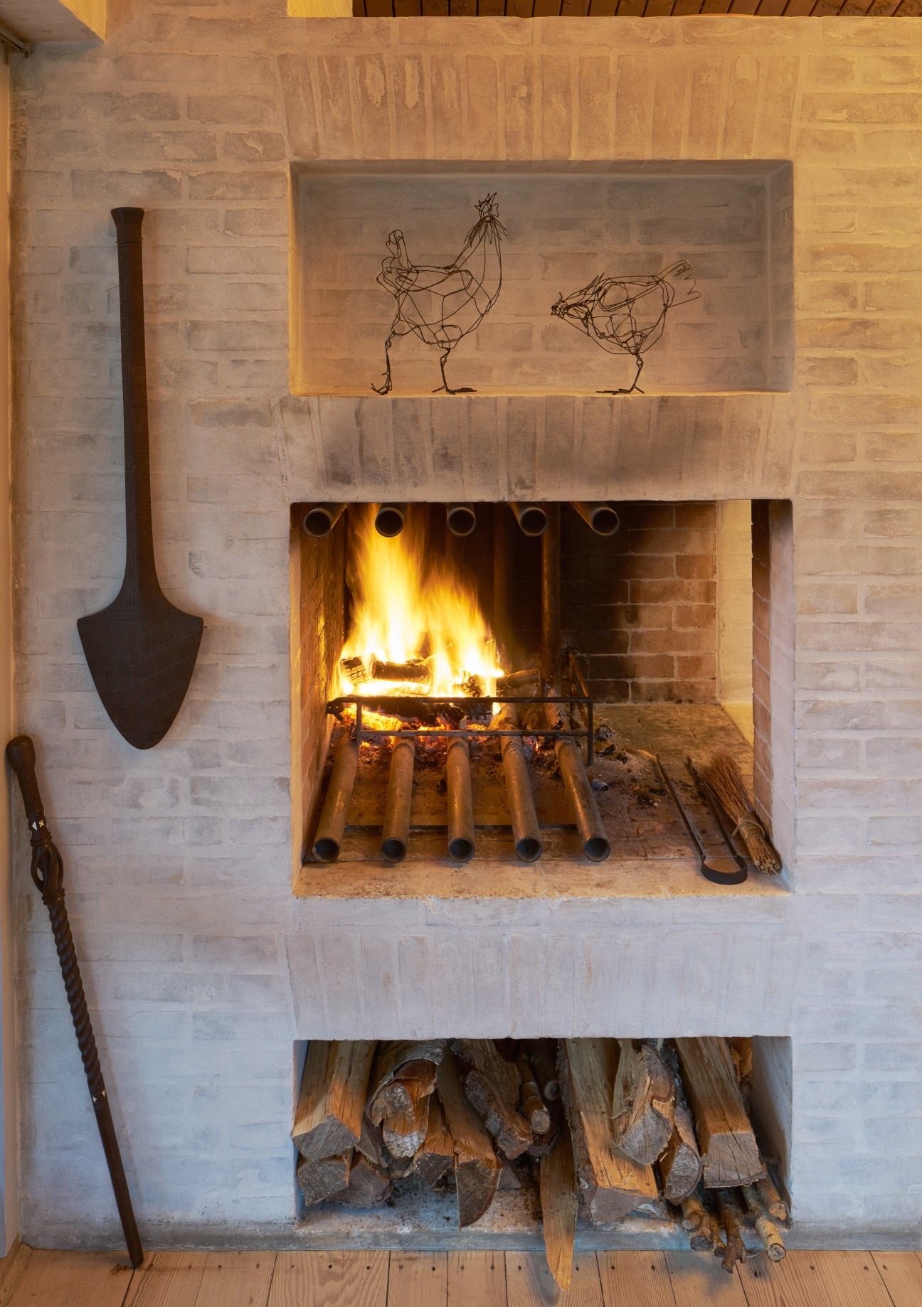 A brick fireplace with a lit fire, metal shovel, wire artwork above, and firewood stored below.