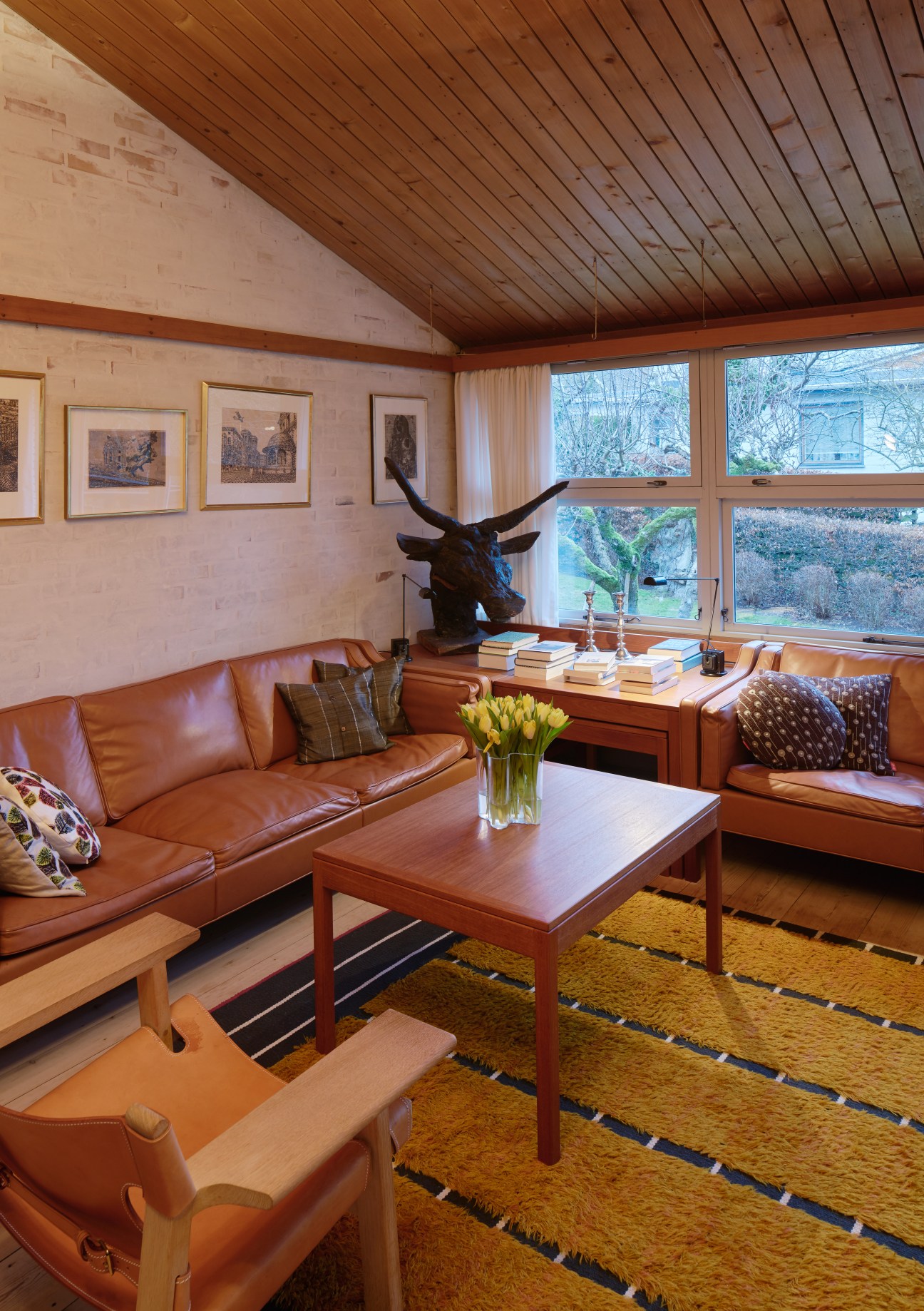 Mid-century modern living room with two cognac leather Mogensen 2213 sofas, wooden desk, and Spanish Chair by large windows.
