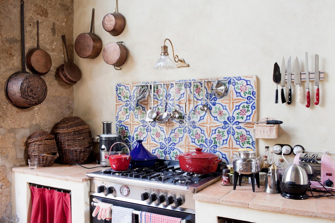Rustic Italian kitchen with copper pans, blue patterned tile backsplash, red cookware, and hanging utensils.