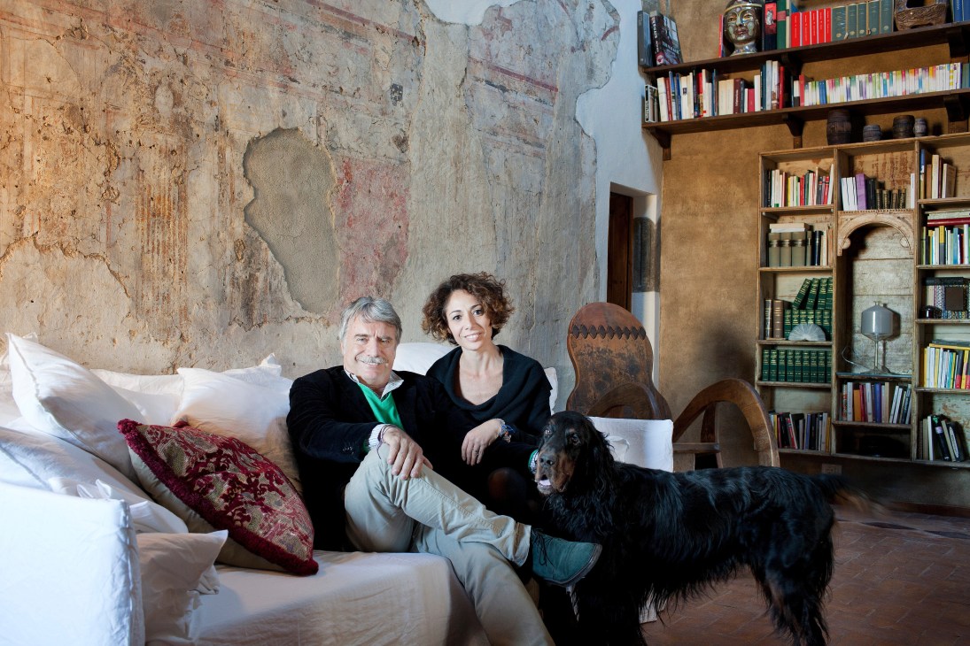 Two people and a dog sitting on a bed in a rustic Italian room with exposed stone walls and bookshelves.