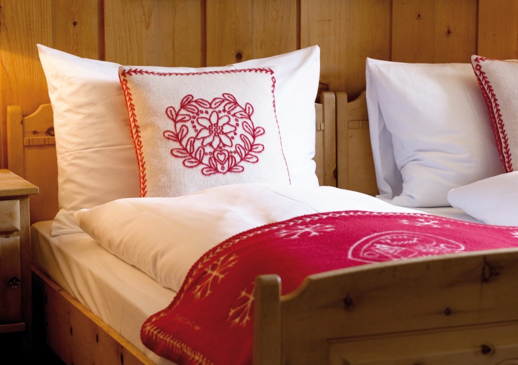 Swiss alpine inn bedroom with wooden walls, white bedding with red embroidered pillows and red patterned blanket.