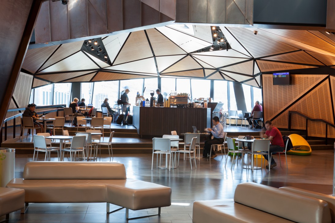 Modern airport terminal interior with geometric ceiling design, seating areas, and coffee counter.