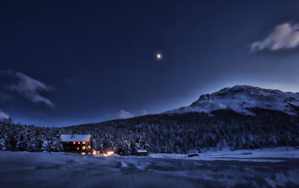 Illuminated Alpine inn beside a frozen lake at dusk with snow-covered mountains and forest under moonlit sky.