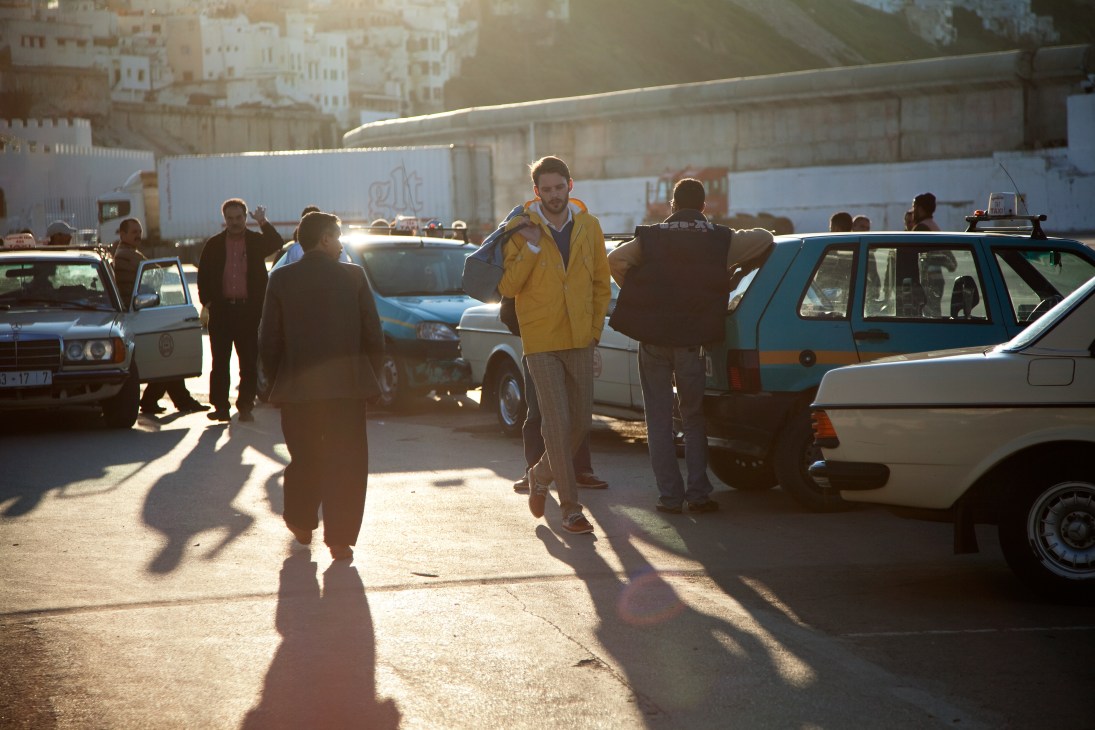 Man in yellow parka stands among parked cars and people in a Tangier street lot.