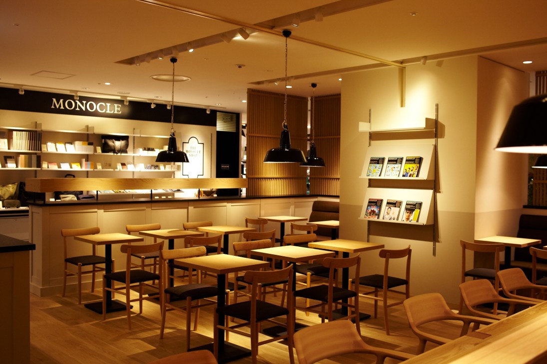 Monocle Café interior with wooden tables, chairs, pendant lamps, shelving, and counter with warm lighting.